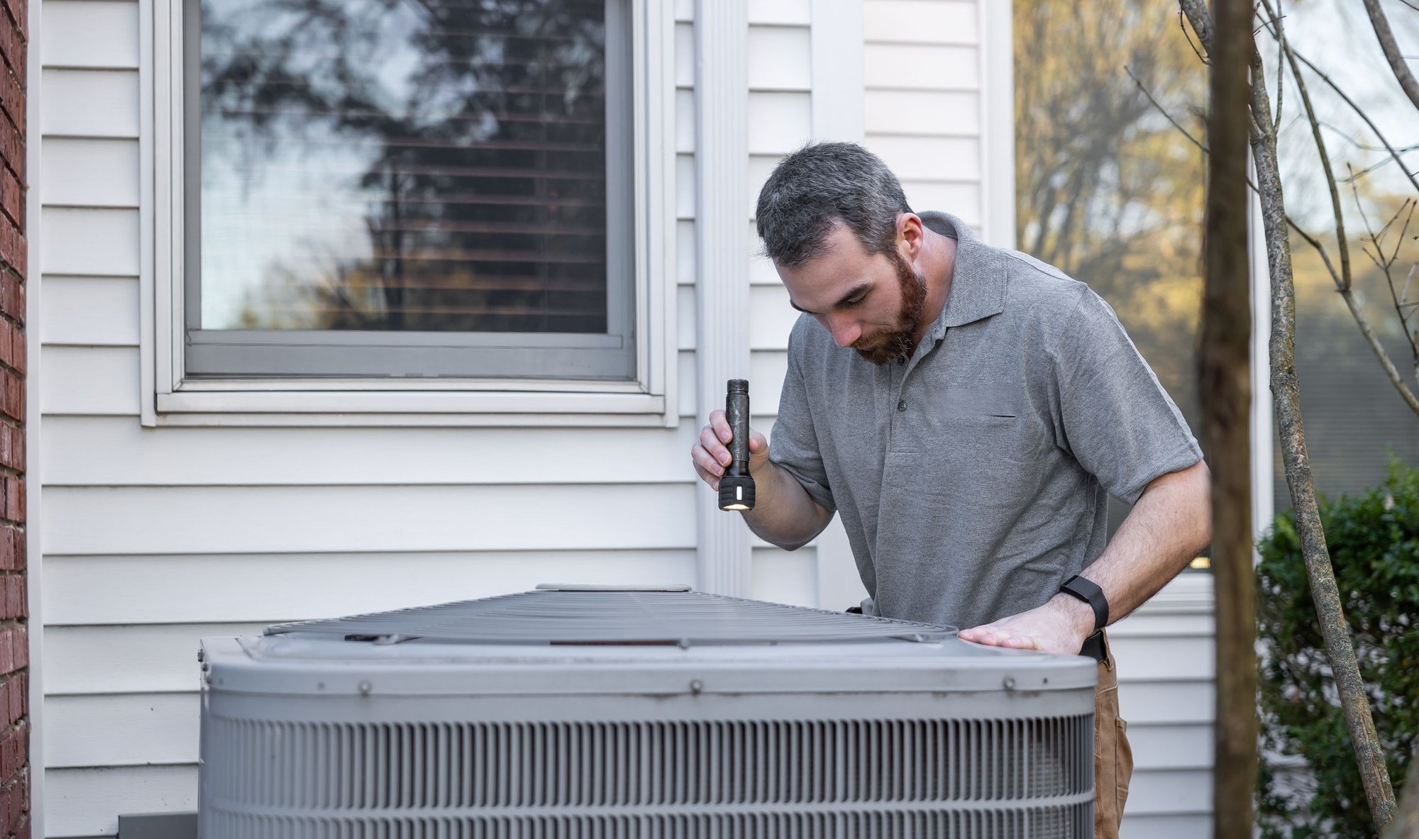 HVAC technician using a flashlight to inspect a home’s outdoor AC unit