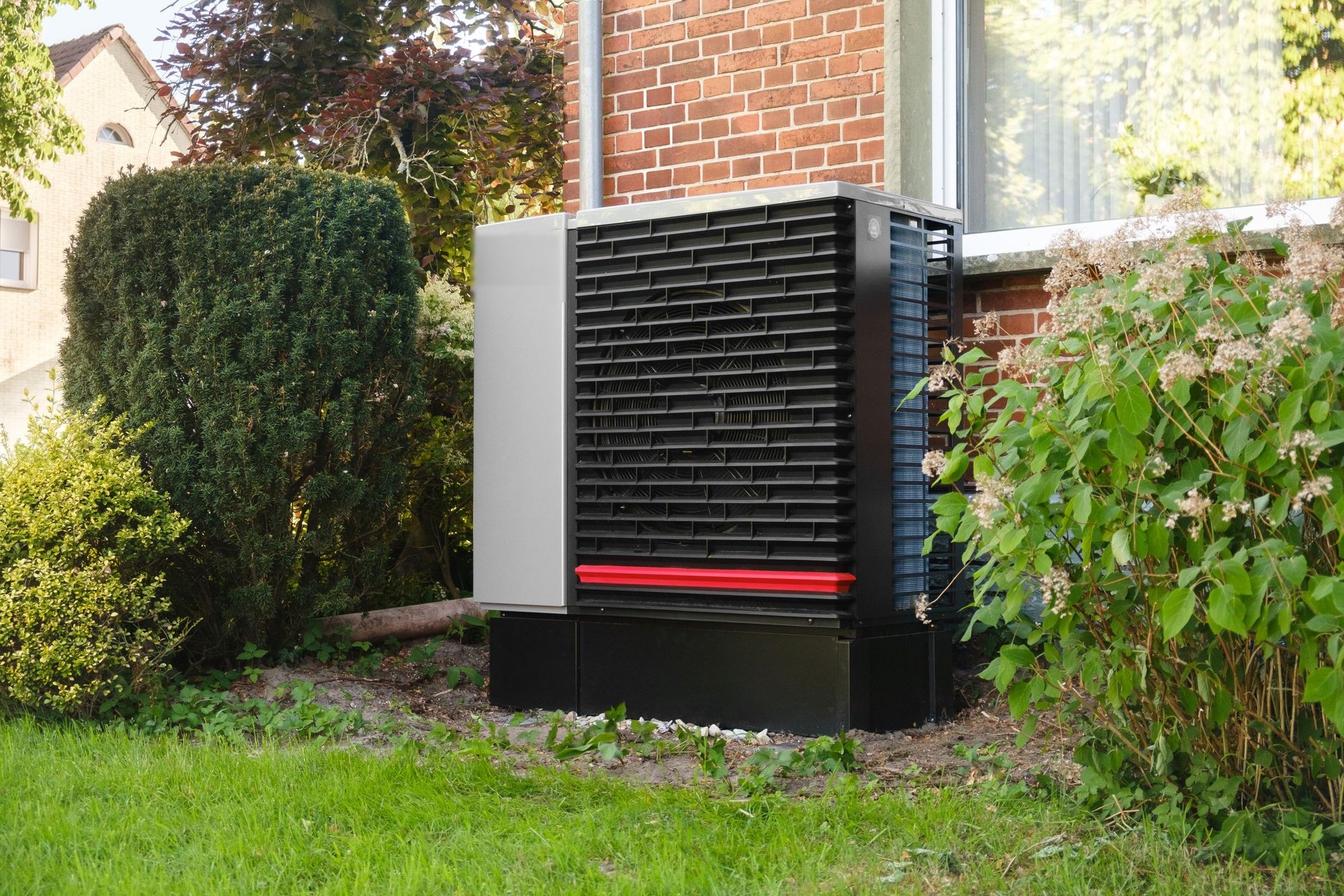 Outdoor heat pump unit beside a brick home surrounded by shrubs and lawn